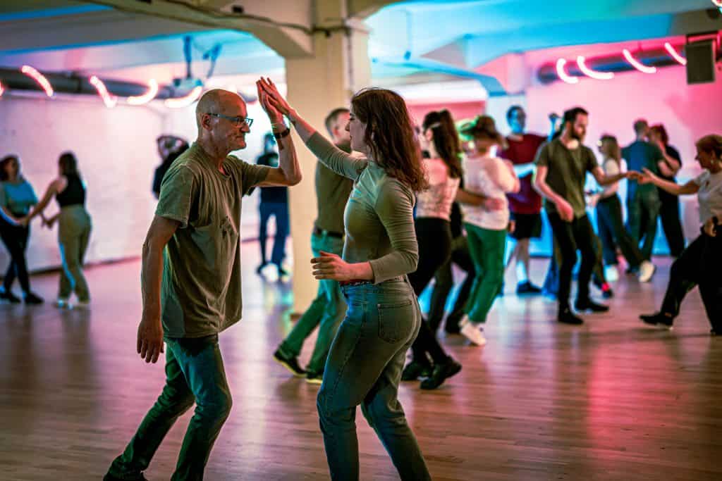 Dancers clap hands overhead while moving on a wooden floor under vibrant pink and blue neon studio lighting.