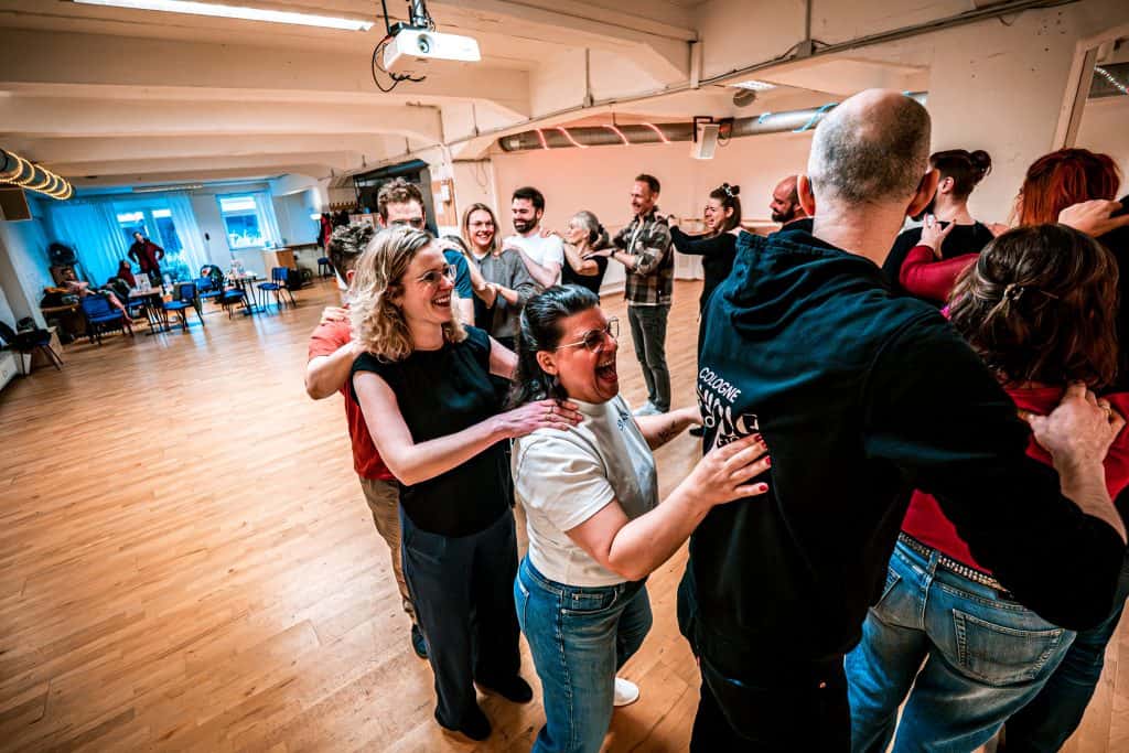 Students in a bright dance studio laugh while forming a train and placing hands on the shoulders of the person in front.