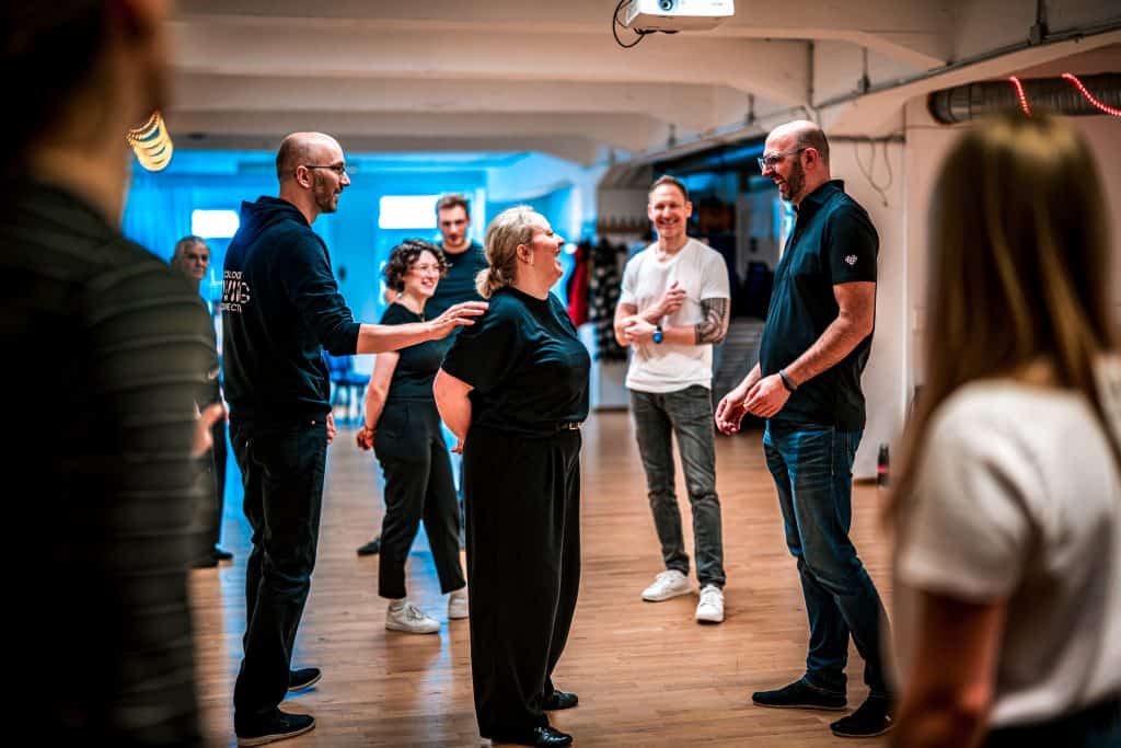 Participants engage in a lively dance lesson while standing on a polished wooden floor in a dimly lit studio.