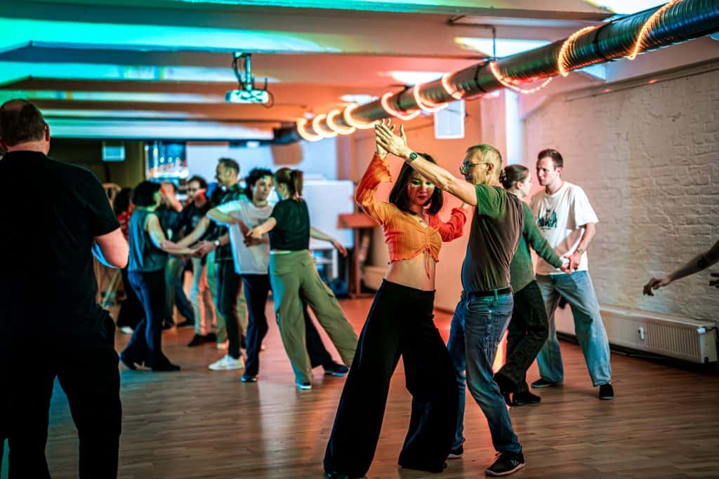 Students practice partner dance moves in a brightly lit studio with teal and orange overhead lighting.