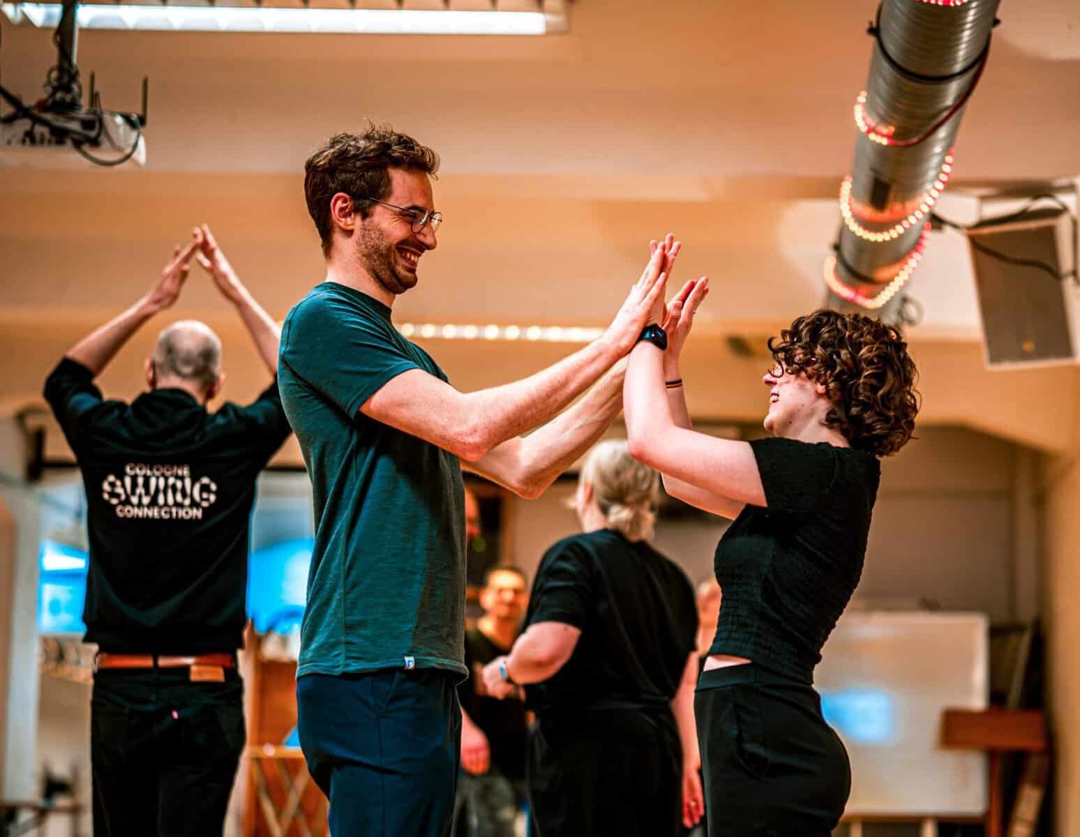 Man and woman laugh while touching palms during a swing dance lesson in a studio with a Cologne Swing Connection logo.