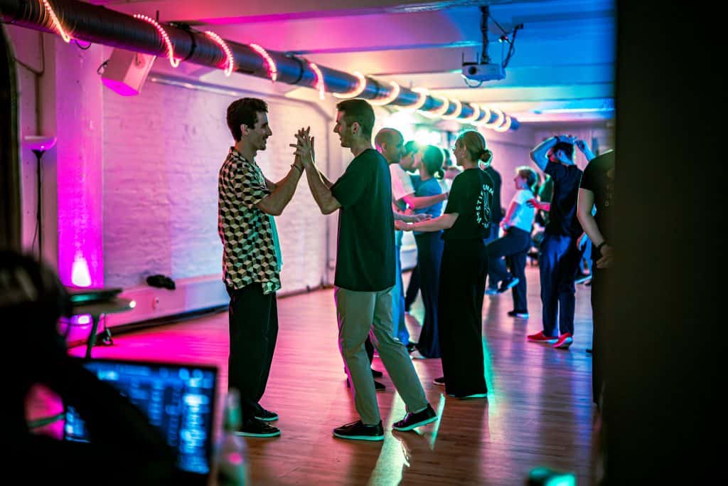 Two men hold hands while dancing in a studio filled with glowing magenta and cyan overhead lights.