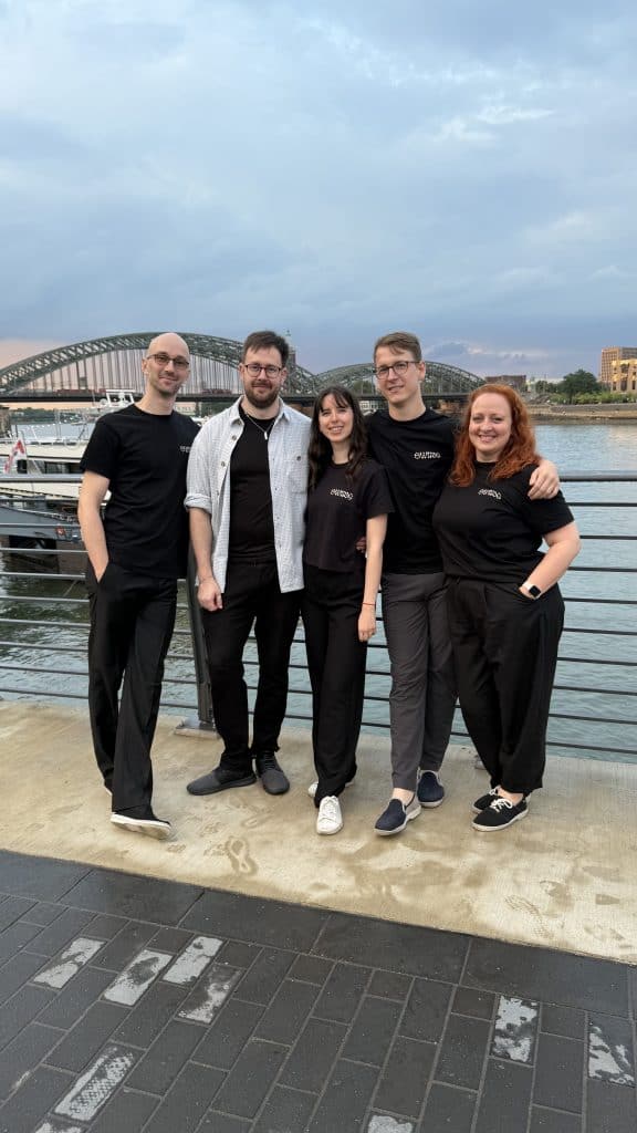 Five people wearing black shirts pose together on a riverside promenade in Cologne with the Hohenzollern Bridge behind.